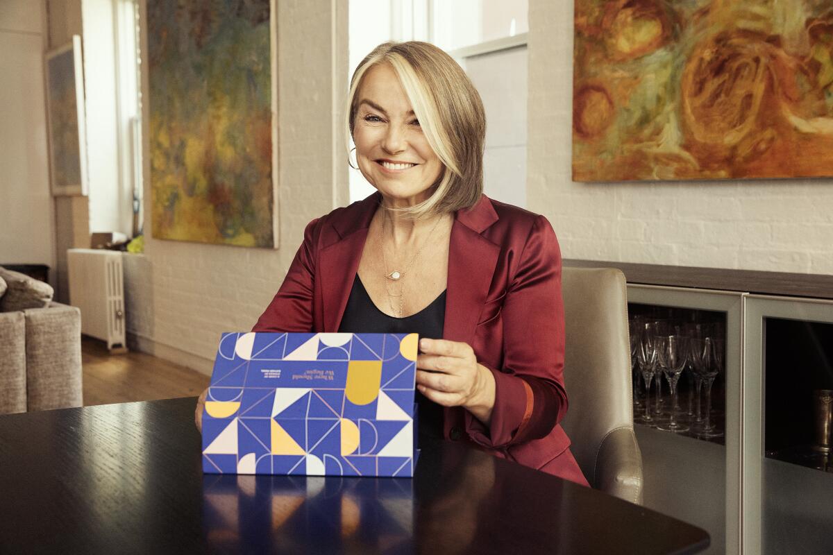 A woman sits at a dining table and smiles while holding up a box