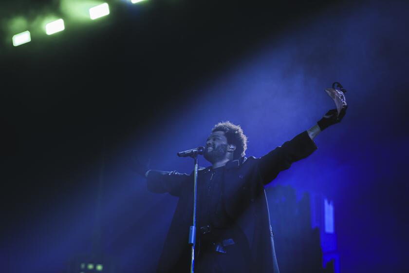 A man with a beard wearing a black coat and outstretching his arms in front of a microphone on a stage with blue lighting