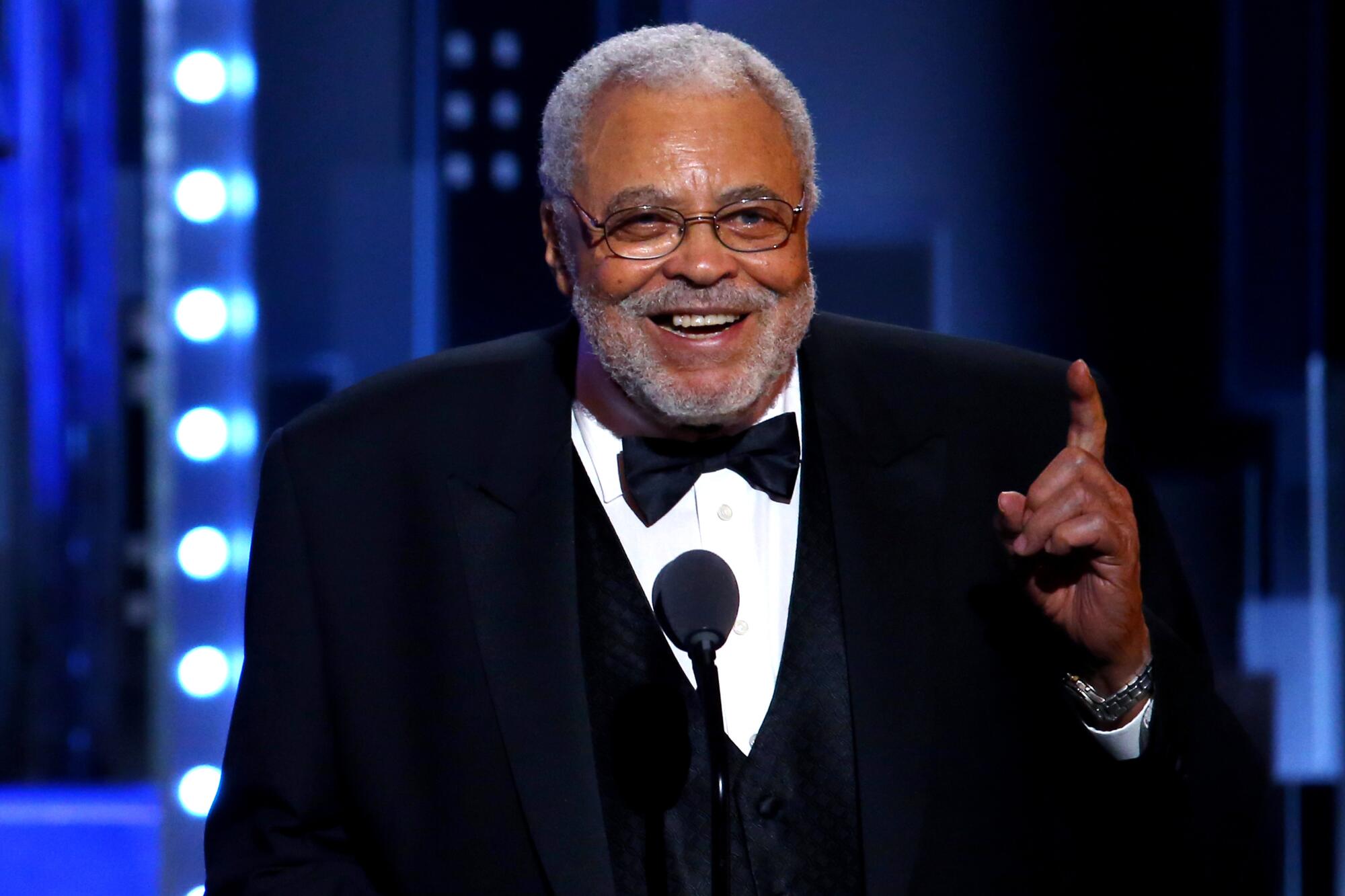 James Earl Jones standing at a microphone in a tuxedo smiling and pointing upward with one hand