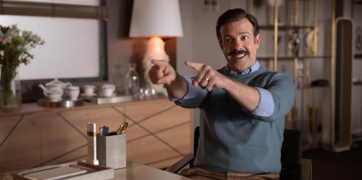 Jason Sudeikis sits at a desk and points with both hands in an episode of 'Ted Lasso.'