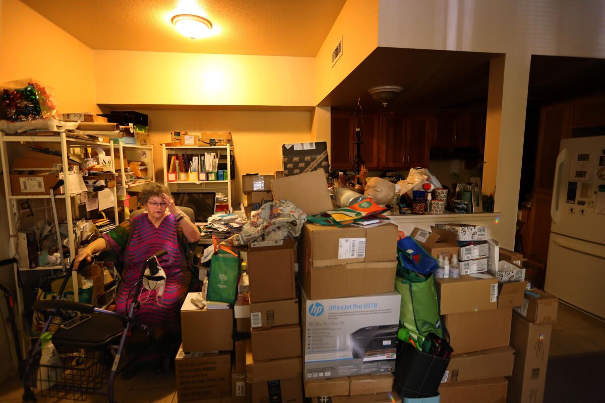 A woman sits in a chair behind her walker, surrounded by boxes and shelves full of belongings
