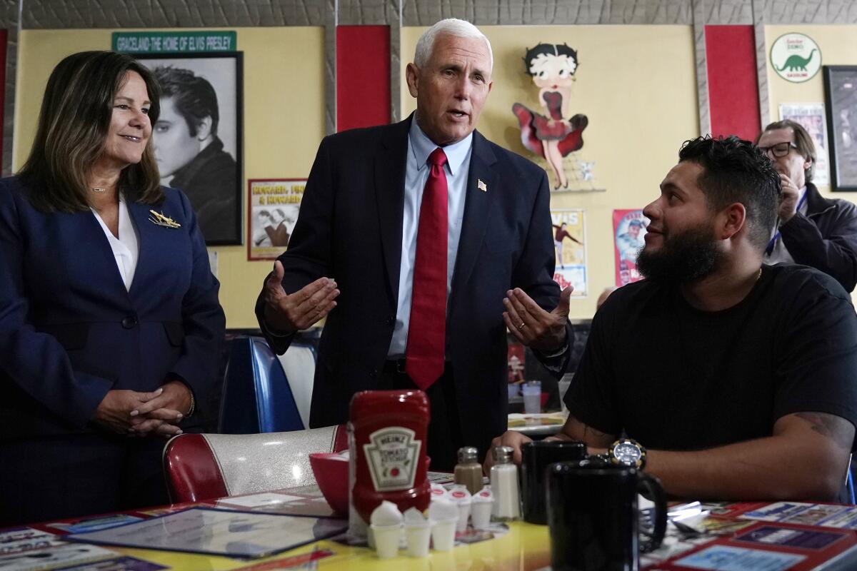 Former Vice President Mike Pence, standing in a diner with wife Karen Pence, gesturing as he talks with a seated patron