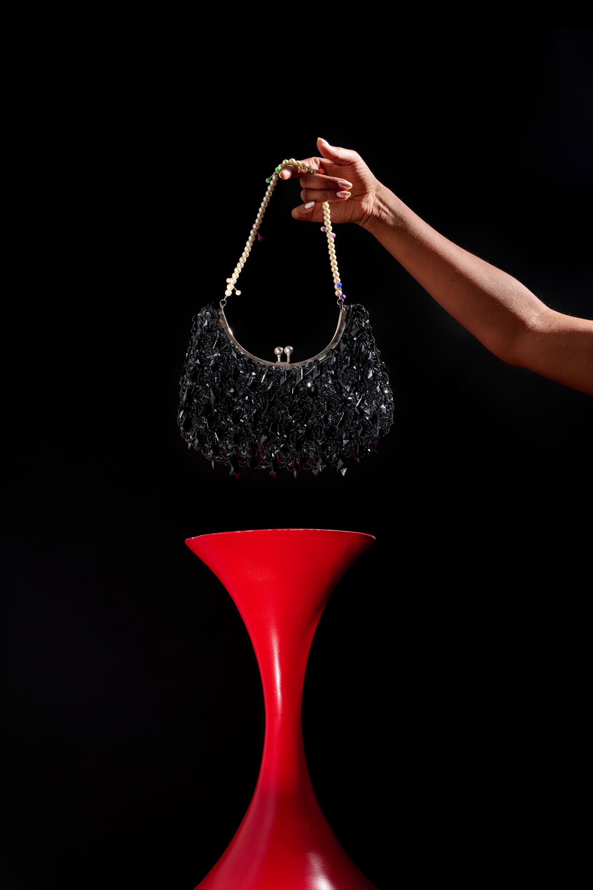 A hand holds a sparkly black purse above a red pedestal