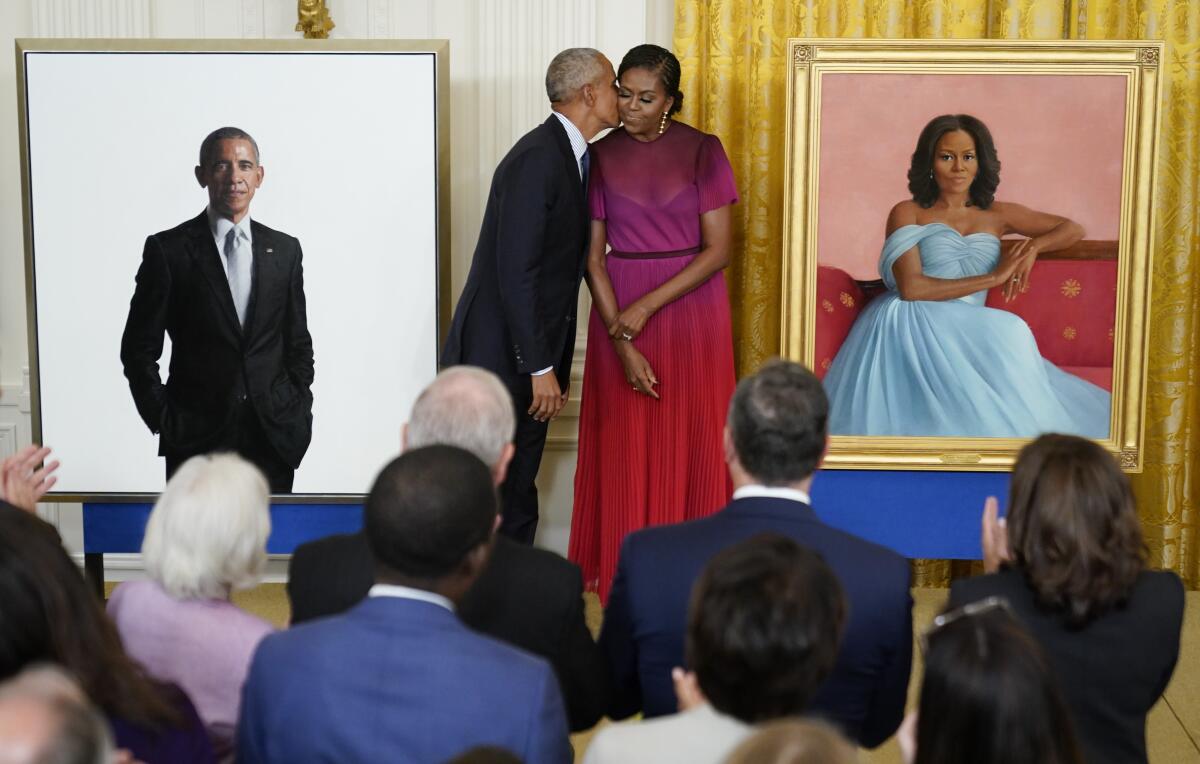 Former President Obama kisses Michelle Obama on the cheek as they stand between their portraits.
