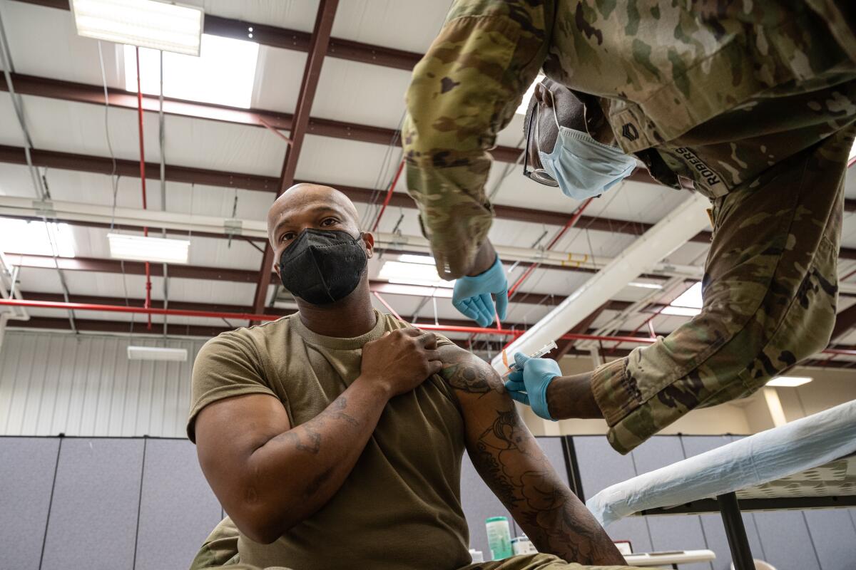 A masked man rolls up his T-shirt sleeve as a masked soldier in uniform sticks a needle in his arm 