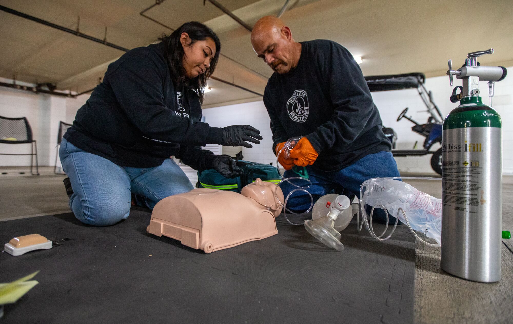 Two people kneel over a training dummy.