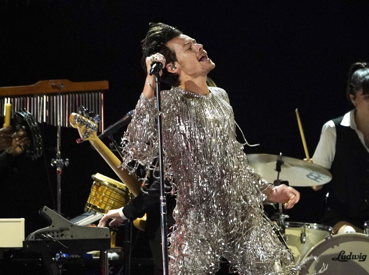 Harry Styles in a sparkly, silver fringe body suit singing on a stage and holding a microphone in his right hand