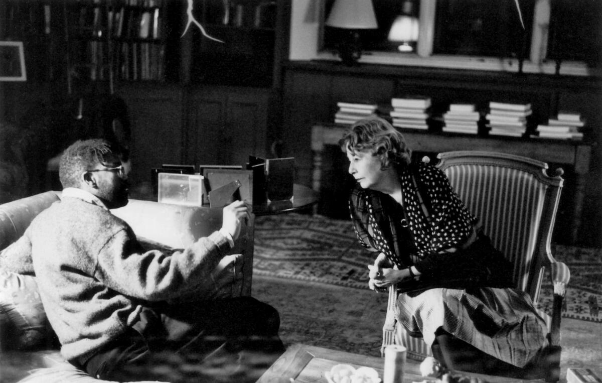 Black and white photo of a young man on a couch, left, talking to a woman in an arm chair