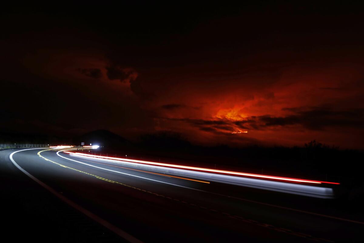 Mauna Loa erupts in the distance behind a road illuminated by headlights.