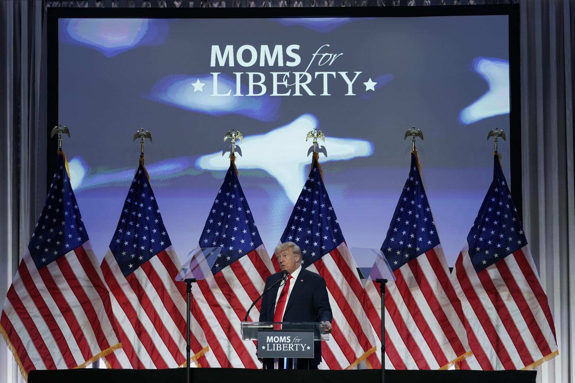 Former President Donald Trump speaking under the words Moms for Liberty and in front of several American flags.