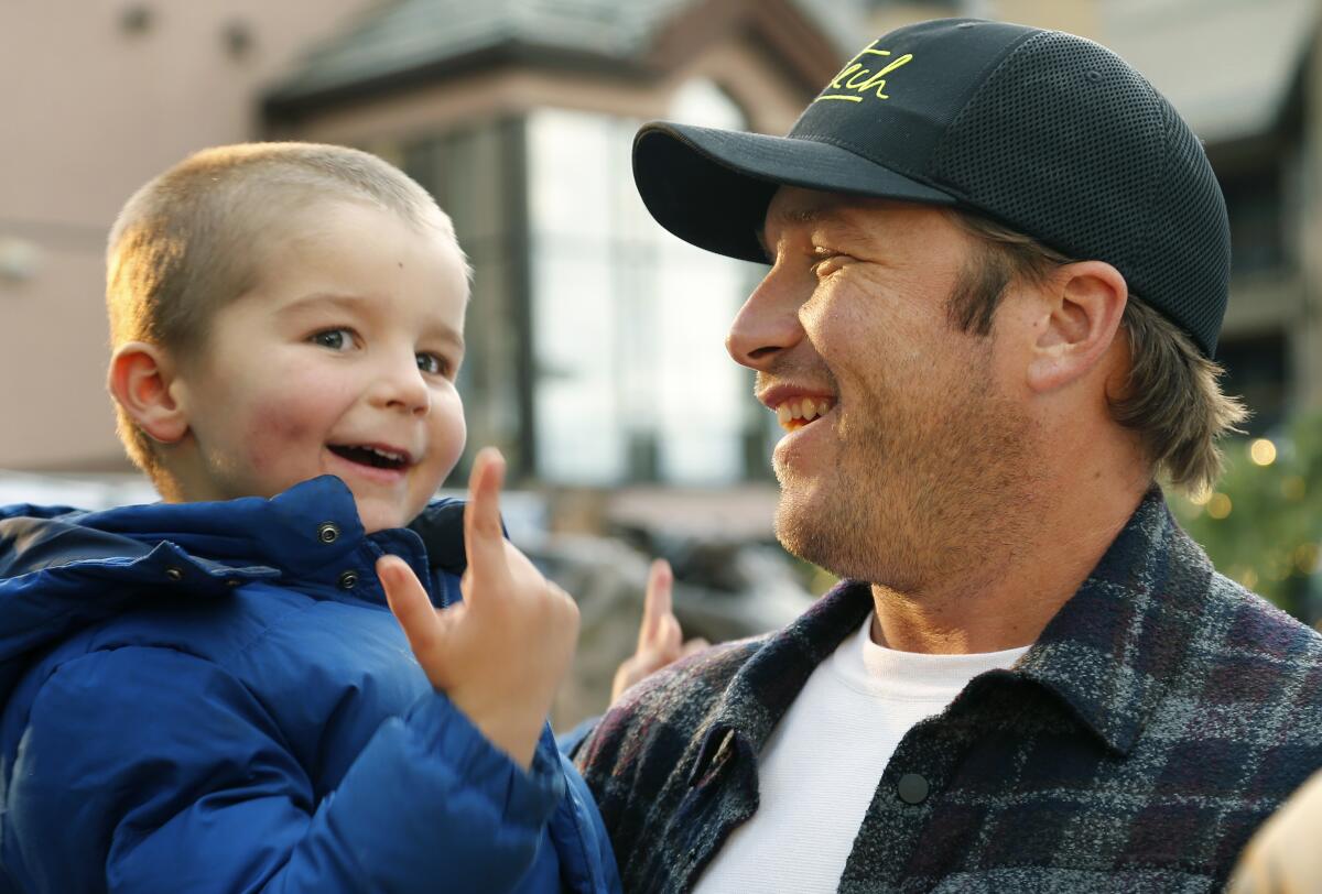 Skier Bode Miller wears a ballcap holds one of his children, who smiles and points up