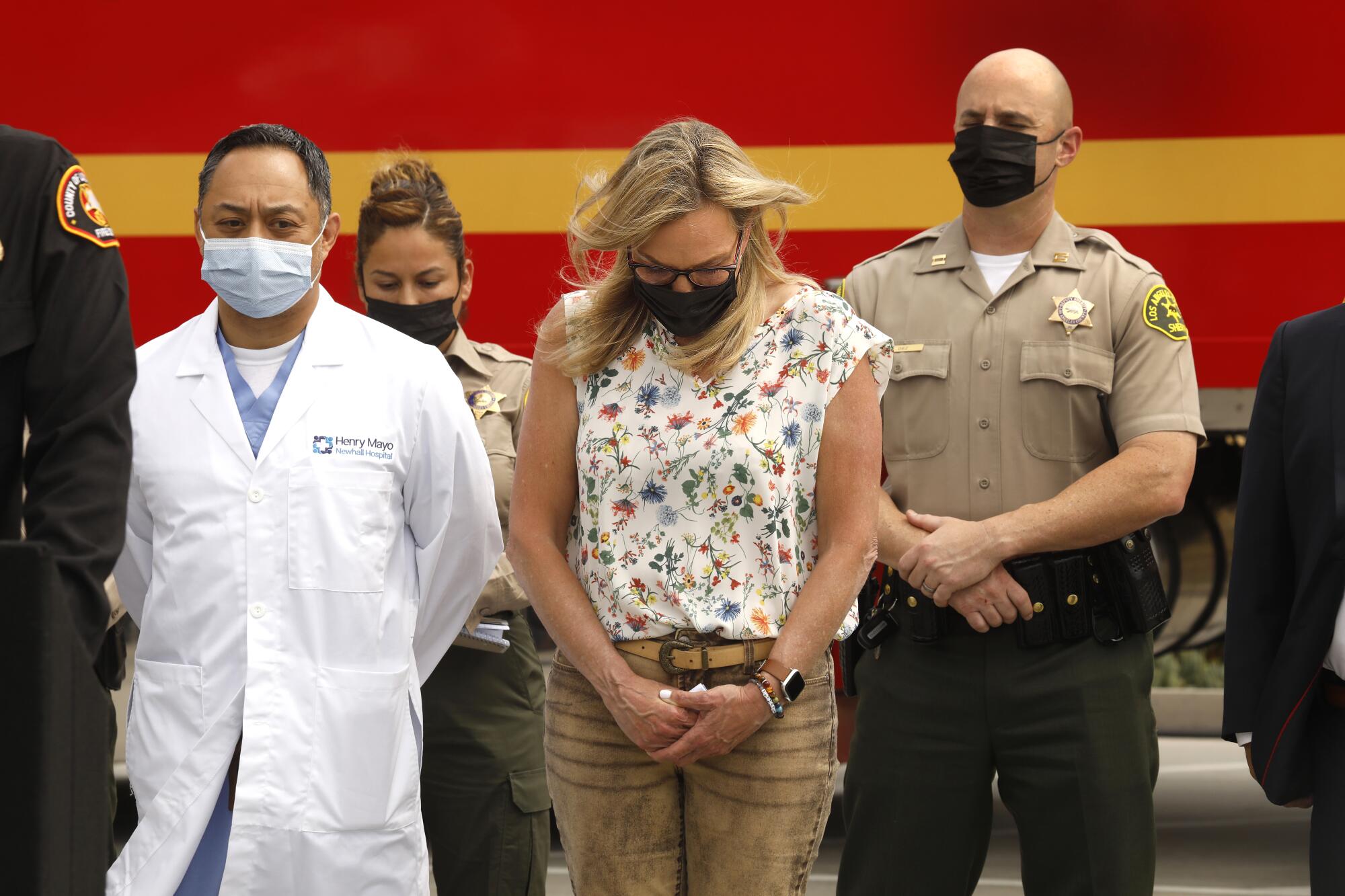 A woman stands outside alongside a doctor in a white coat and two deputies. They all bow their heads.