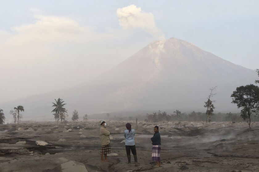 Villagers stand on an area covered in volcanic ash as Mount Semeru looms in the background in Kajar Kuning village in Lumajang, East Java, Indonesia, Monday, Dec. 5, 2022. Indonesia's highest volcano on its most densely populated island released searing gas clouds and rivers of lava Sunday in its latest eruption. (AP Photo/Imanuel Yoga)