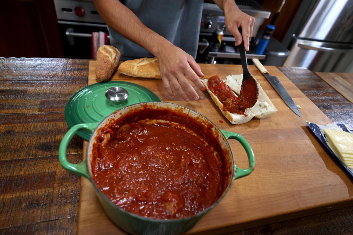 Han spoons tomato sauce from a large green pot onto a cut sandwich roll