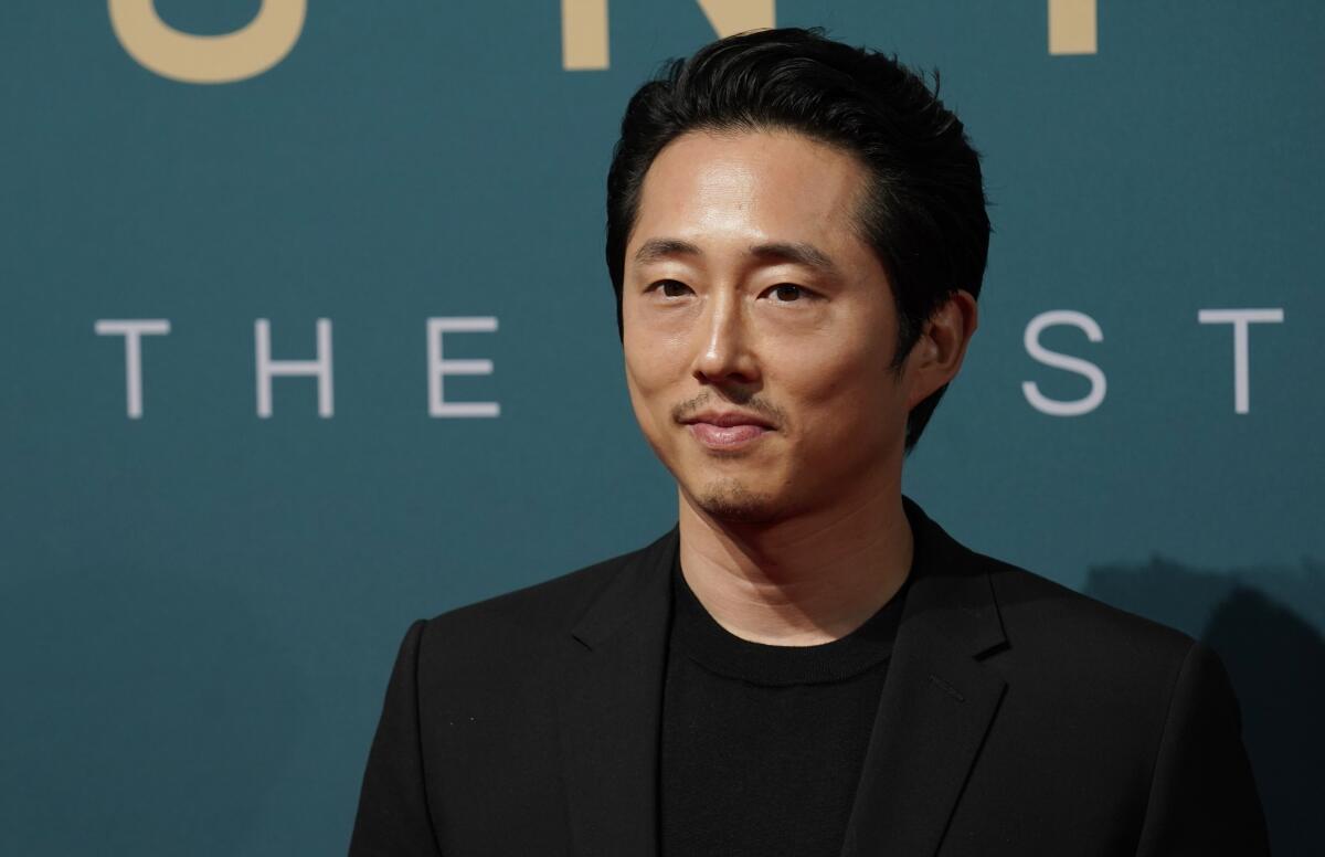 Steven Yeun stands with a slight smile, his mouth closed, wearing a black suit jacket and black T-shirt