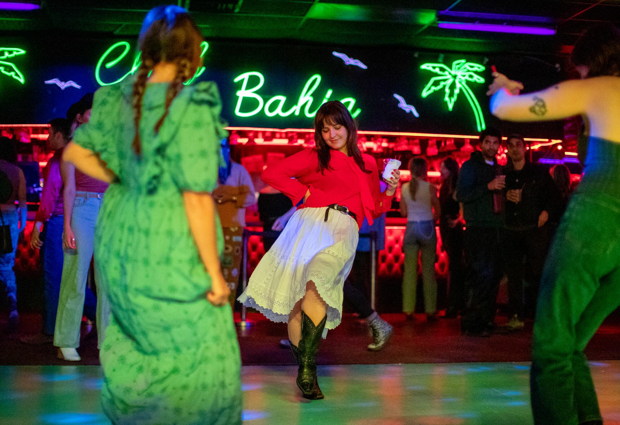 A person in a red sweater and white skirt dances on a multicolored dance floor. 
