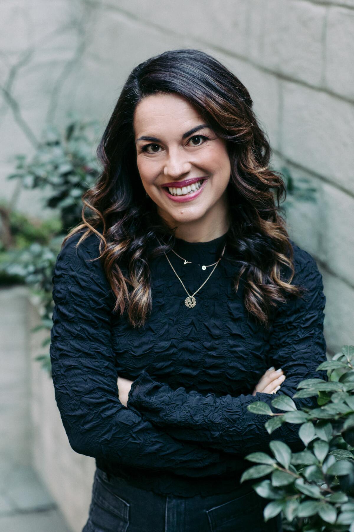 A smiling woman dressed in dark clothing stands with her arms crossed.