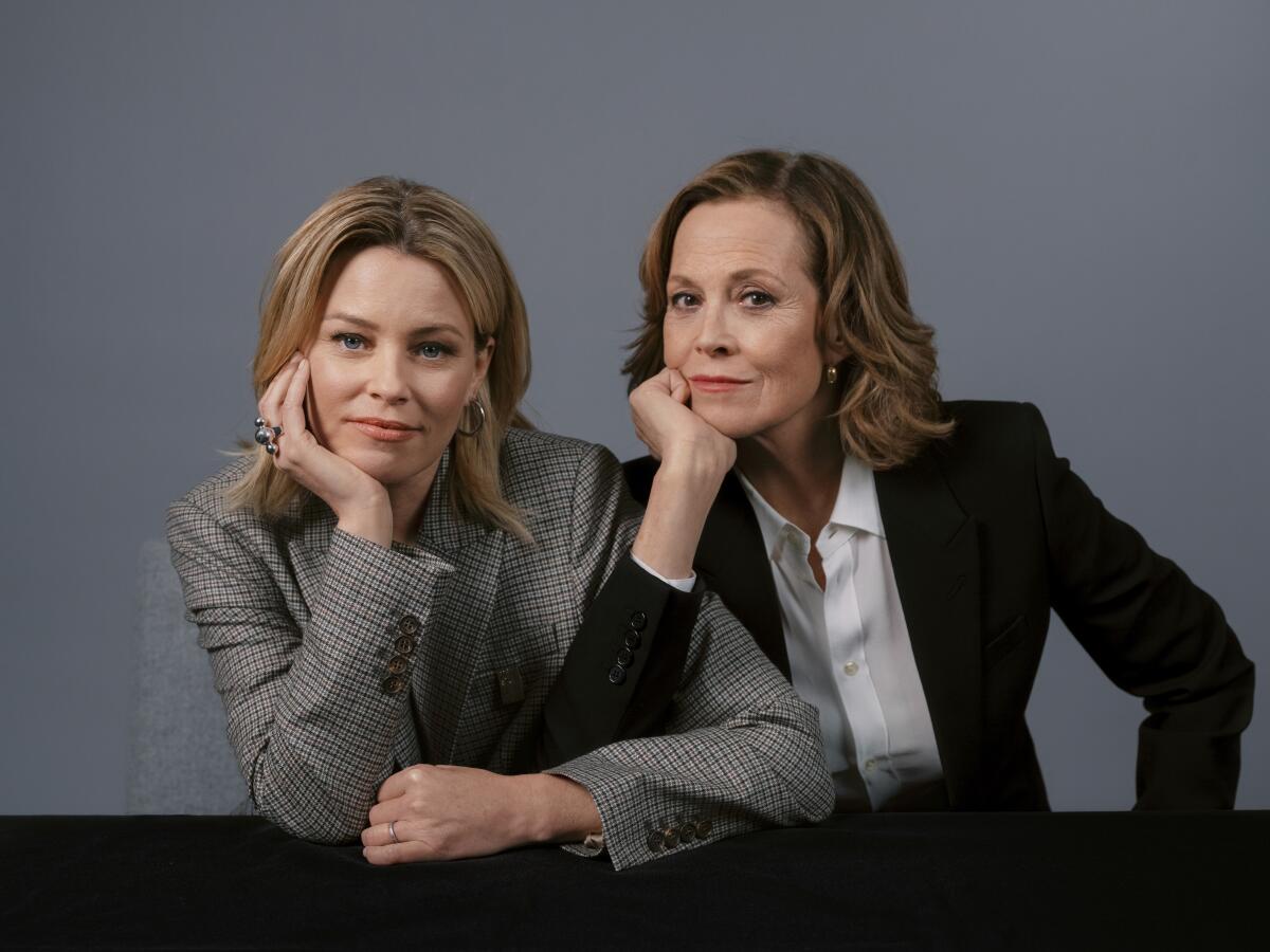 Elizabeth Banks and Sigourney Weaver link arms and rest their chins on their hands for a portrait.