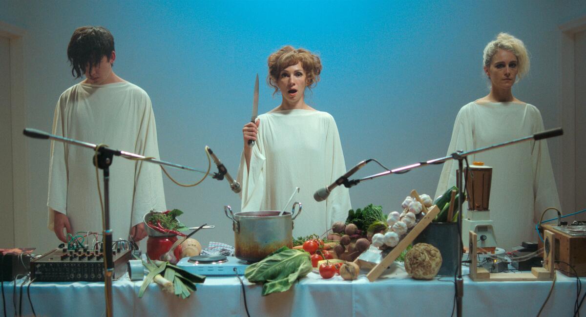 Three people in white robes stand behind a table covered with food.