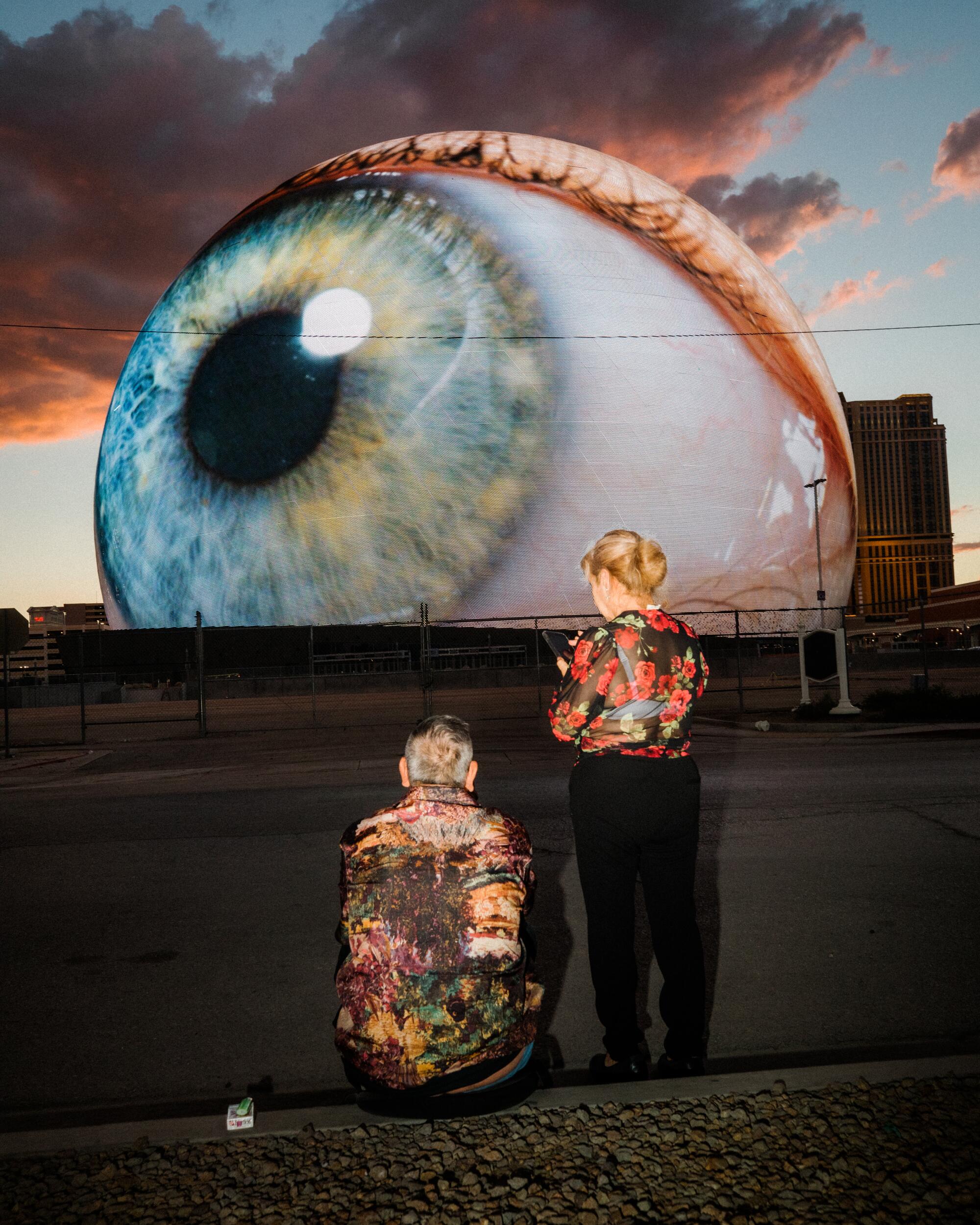 Two people wearing bright, shiny shirts stand observing a spherical building broadcasting an eyeball.