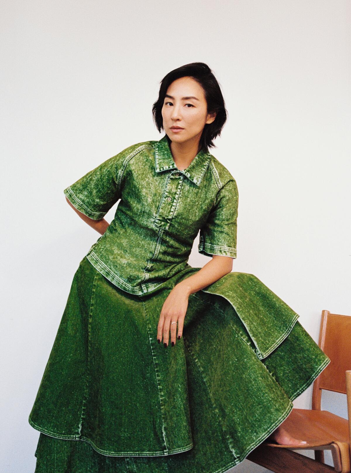 Greta Lee rests one foot on a chair, leaning onto that raised leg for a portrait.