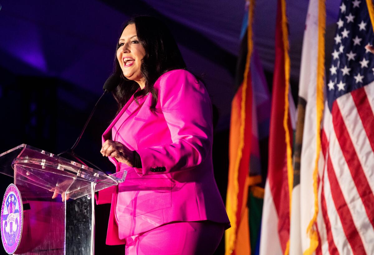 A woman in a bright pink suit speaks at a podium in front of a row of flags
