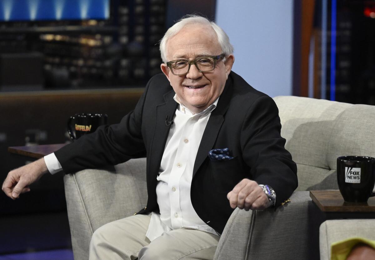 A man with white hair and glasses and a black sport coat sits in gray armchair on a TV set