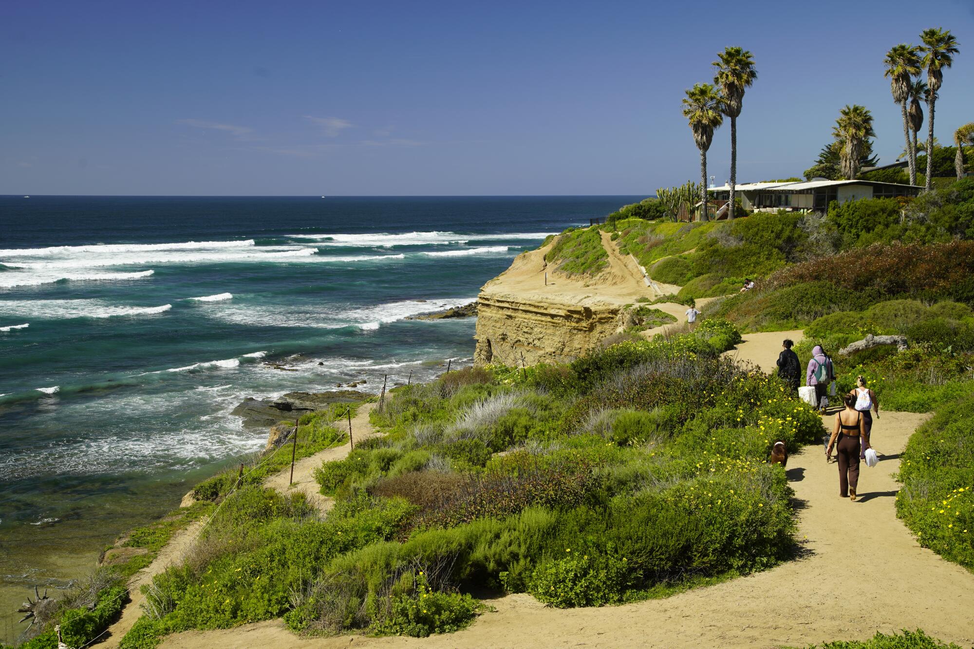 People walk along a sandy path atop verdant cliffs above ocean waves.