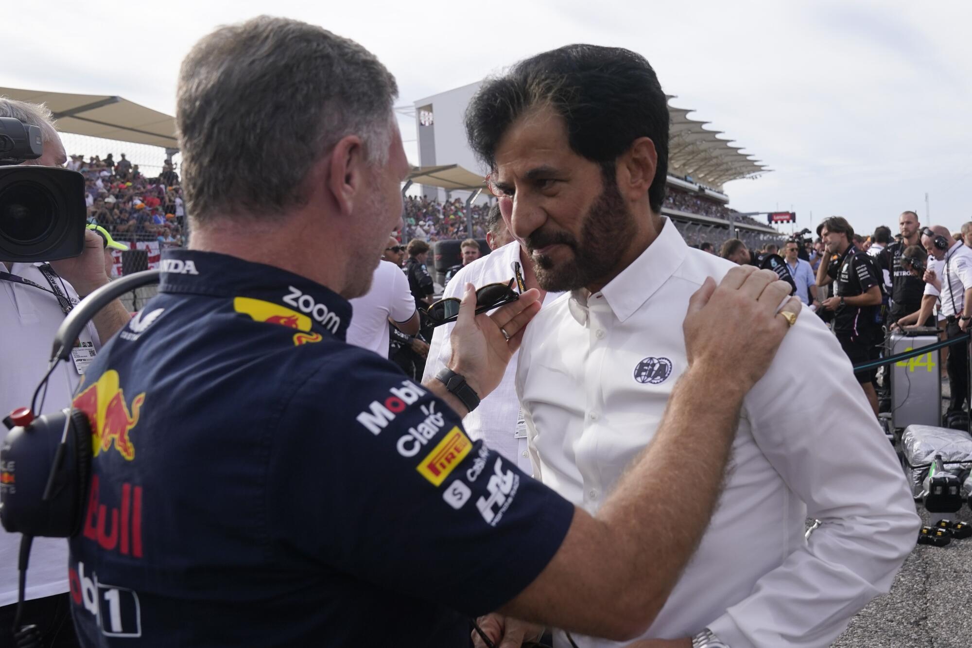 A man wearing race team clothing speaks to a man wearing a white buttondown with a logo that reads "FIA." 