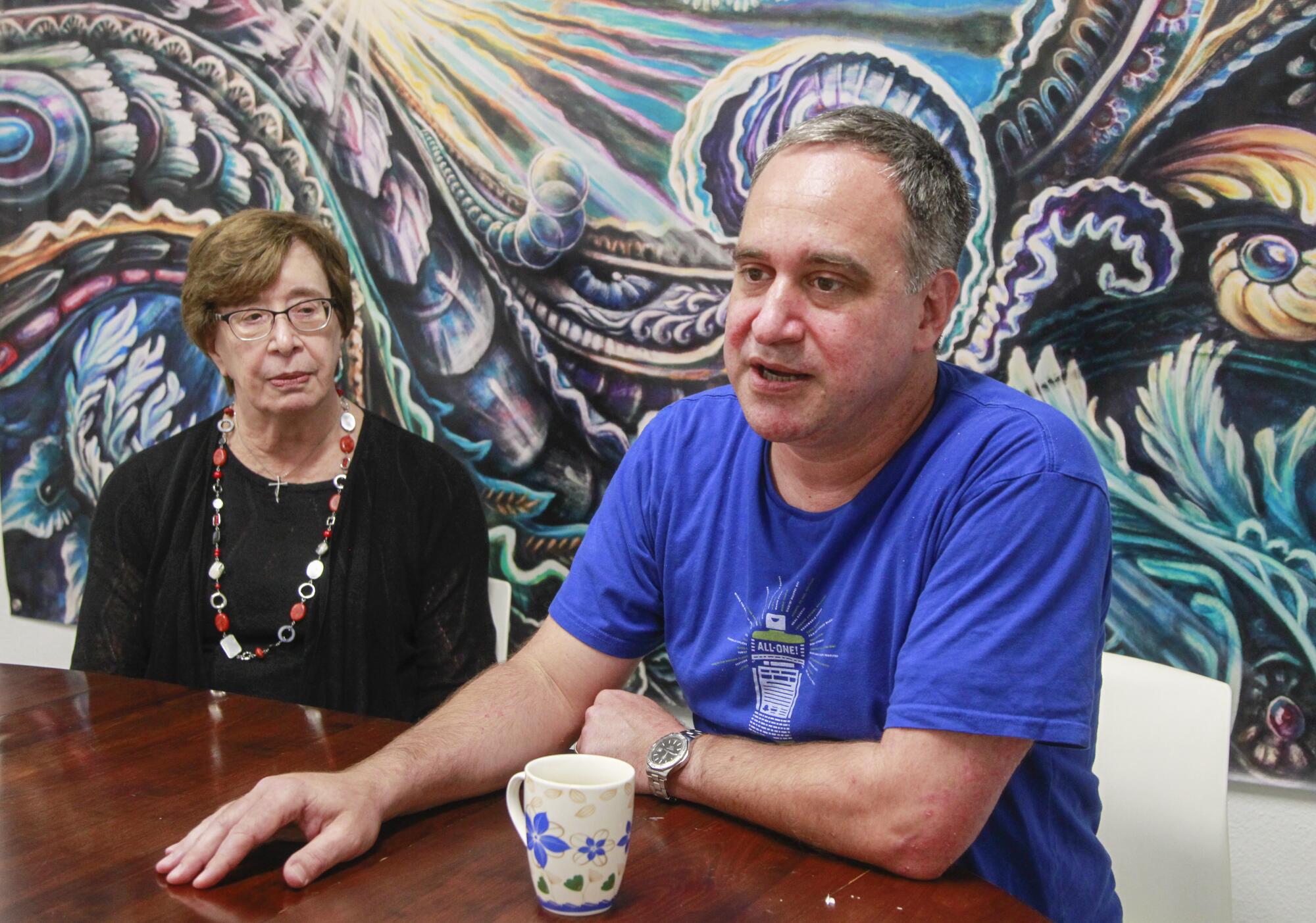 A woman sitting at a table beside a man as he speaks, against a backdrop of a wall with a fanciful psychedelic design.