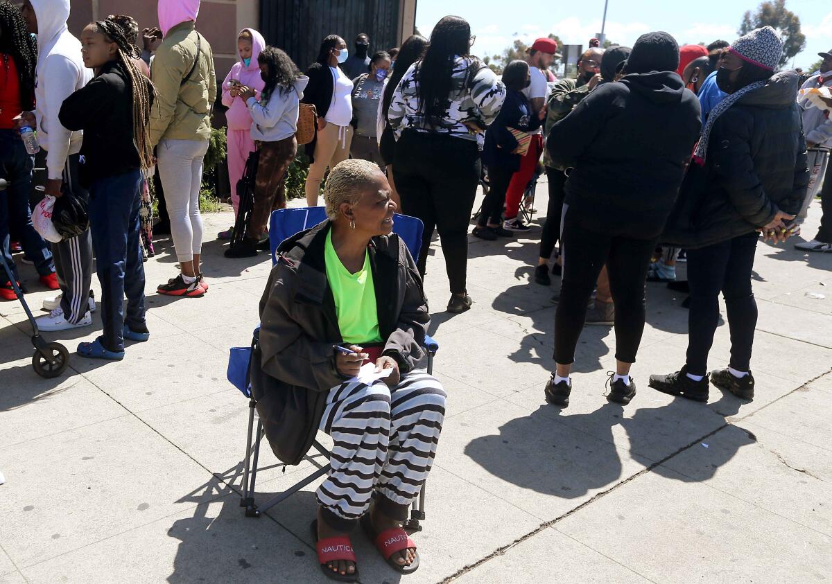 A woman sits in a folding chair next to a line of people on a sidewalk.