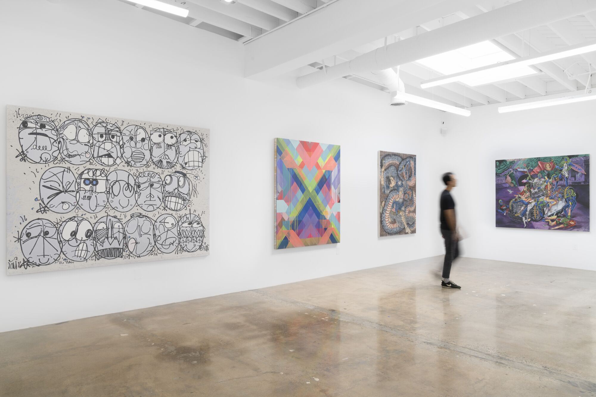 A blurred figure walks past four works of art hanging on the white walls of an open gallery.