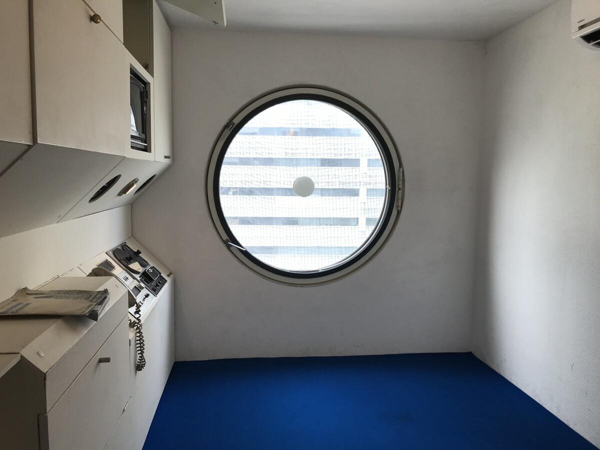 A tiny room with blue carpet, a circular window and built-in drawers and cubby for a TV.