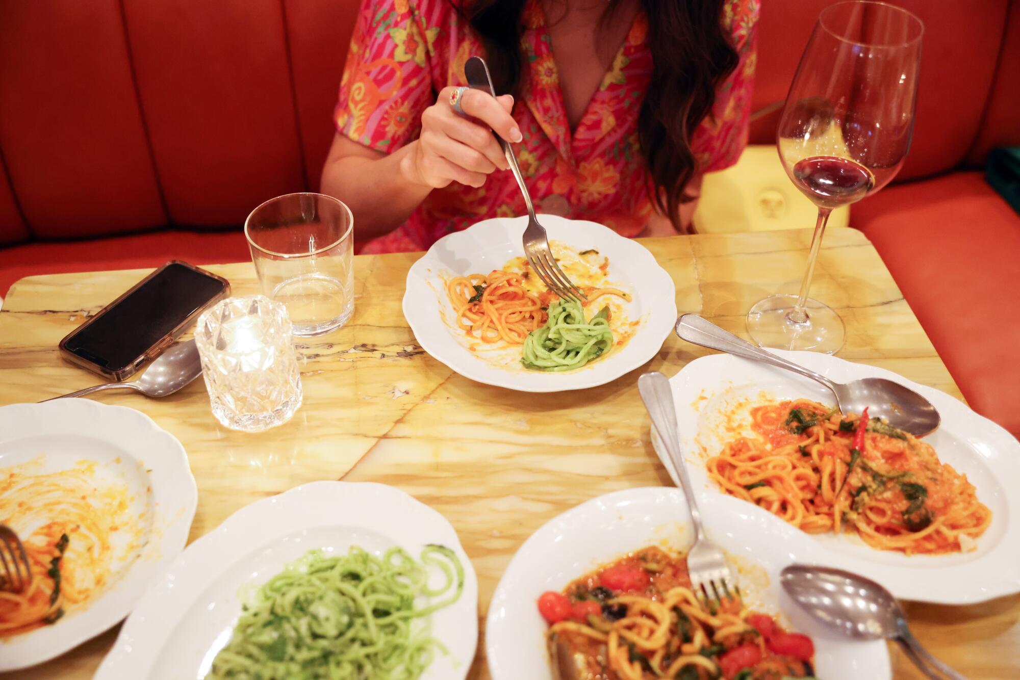 A woman sticks a fork into one of several plates of different kinds of pasta on the table in front of her.