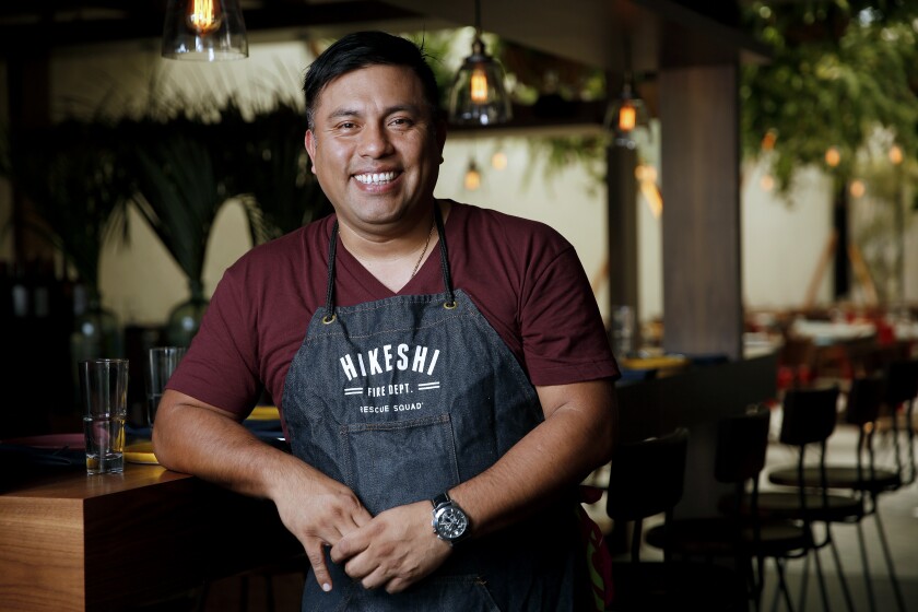 A smiling man in an apron stands in the dining area of a restaurant.