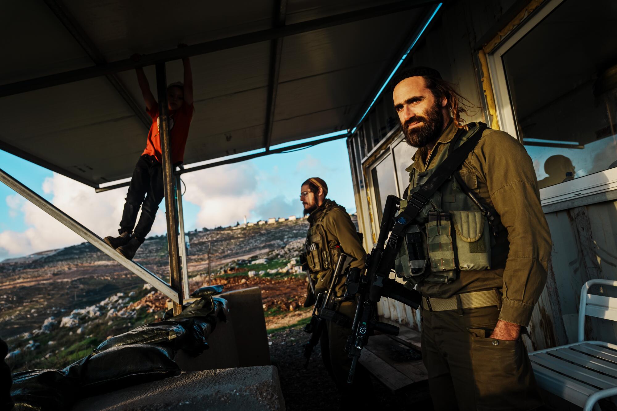 Three soldiers, one propped up on a roof brace, outside a small building, gazing down on communities in the valley below