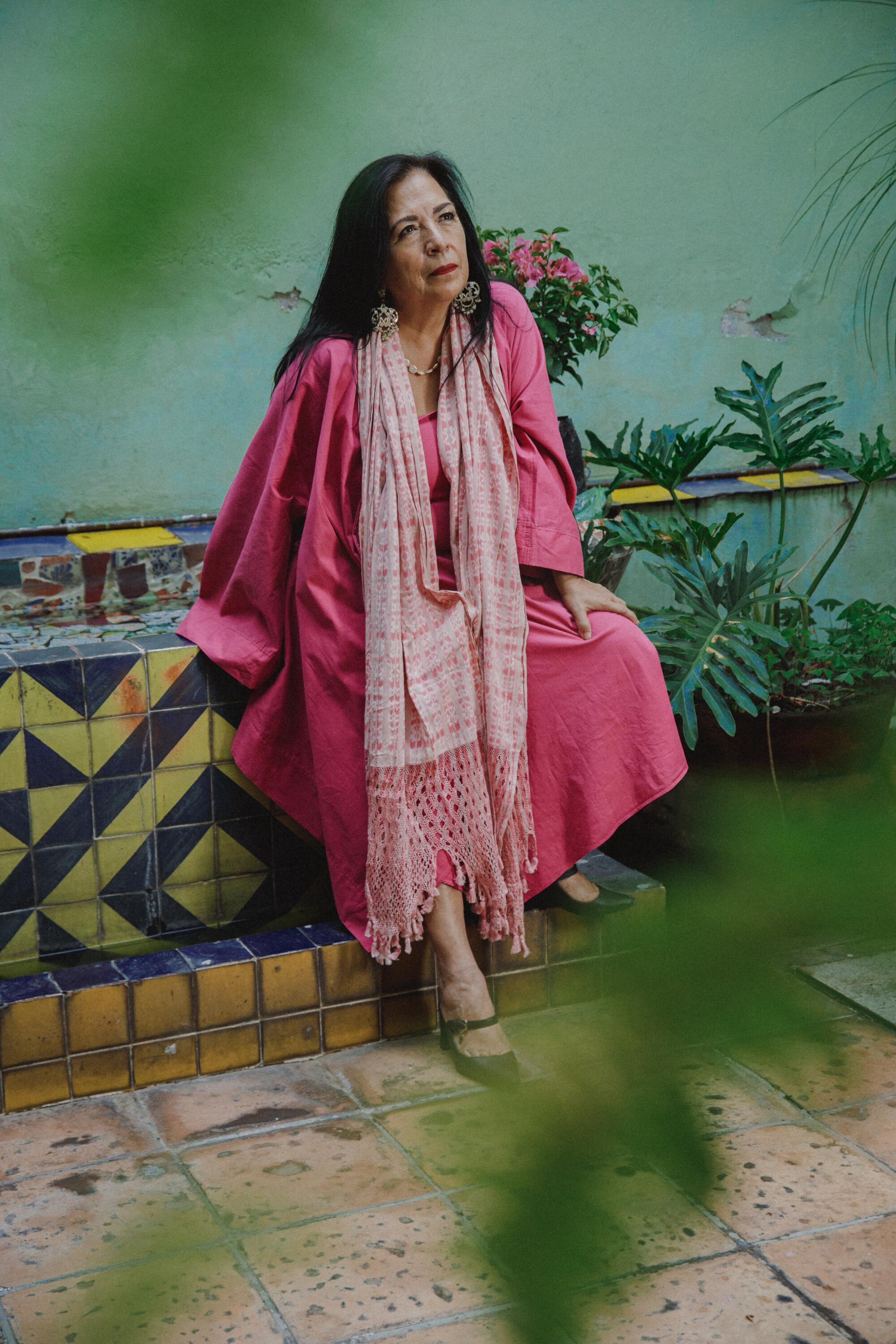 A woman in a long, flowing pink robe sits in a garden.