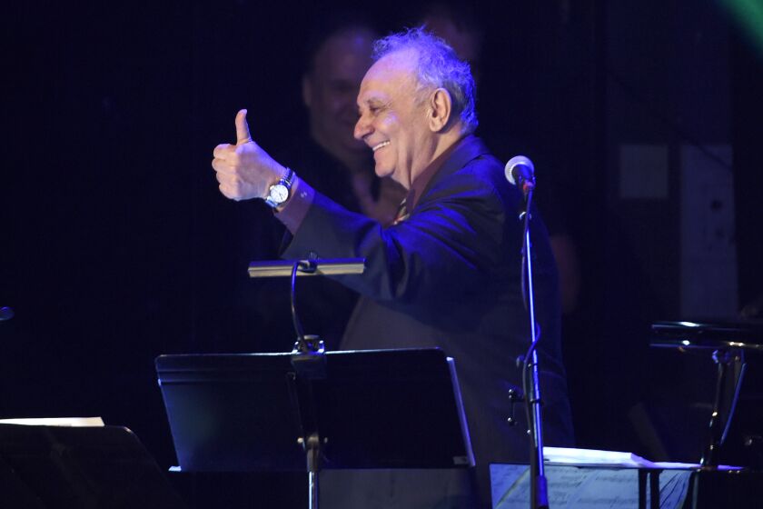 An older, smiling man looks to the side and gives the thumbs-up sign during a musical performance