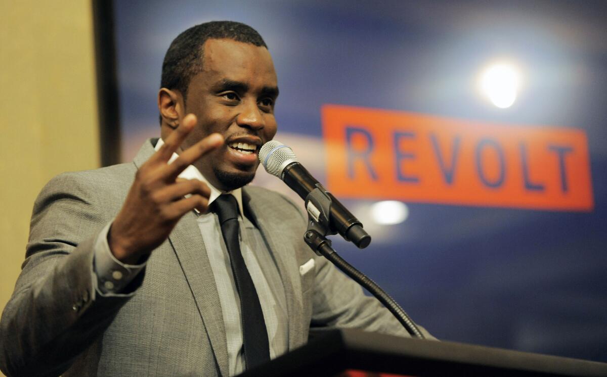 Sean "Diddy" Combs lifts up one hand as he speaks at a mic in front of a Revolt logo while clad in a tan suit and dark tie