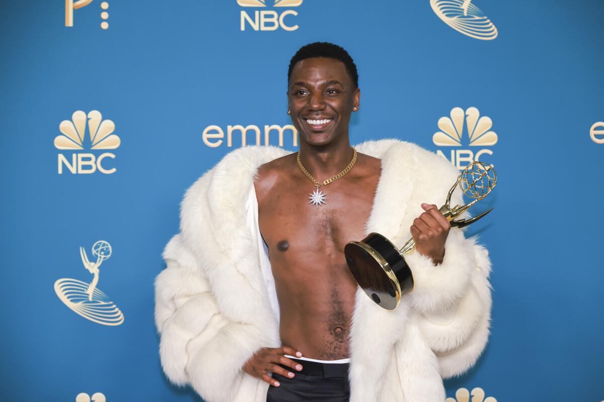 A man wearing a furry white coat draped around his shoulders smiles and holds an Emmy Award