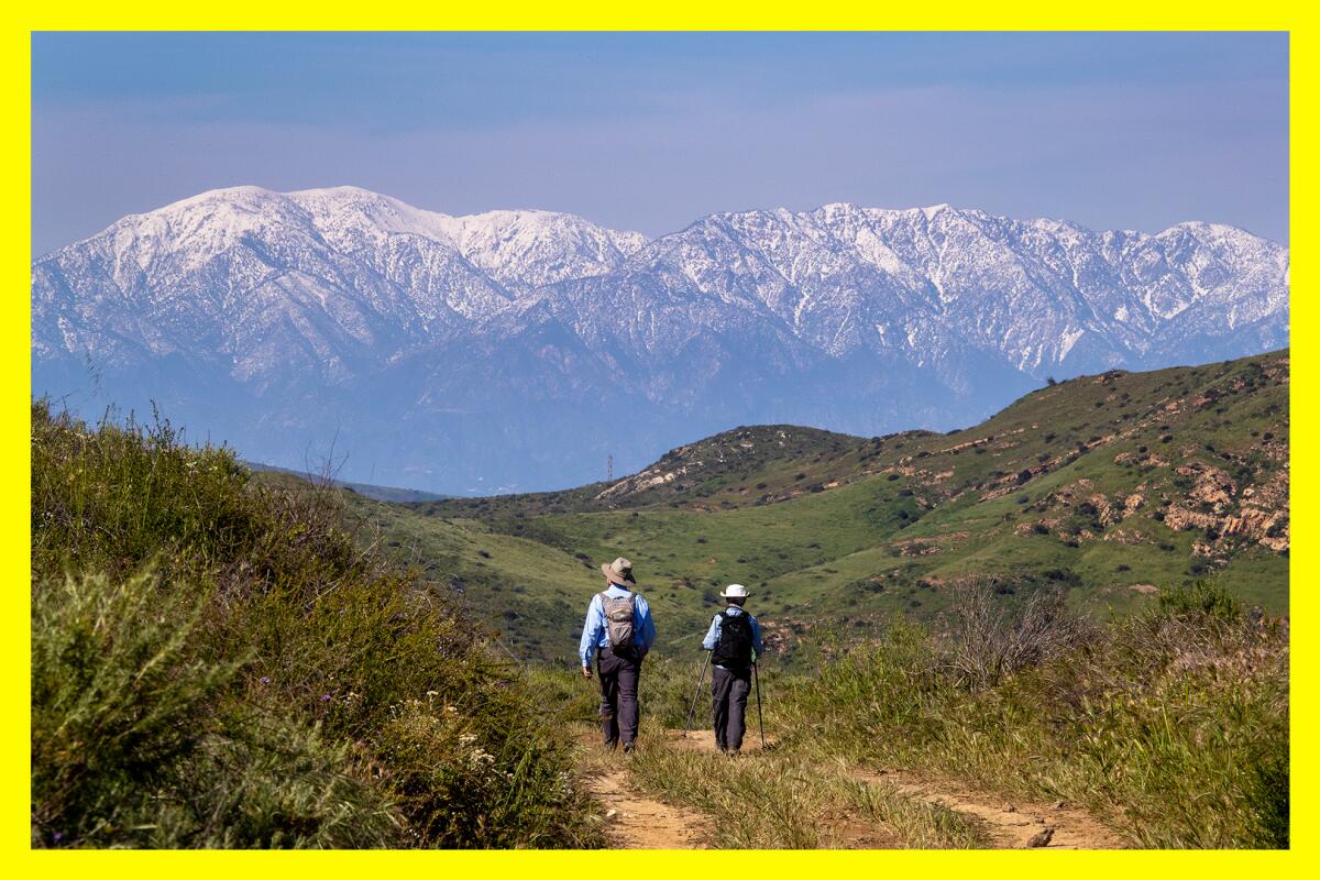 Two hikers on a scenic trail walk toward the snow-capped San Gabriel Mountains.