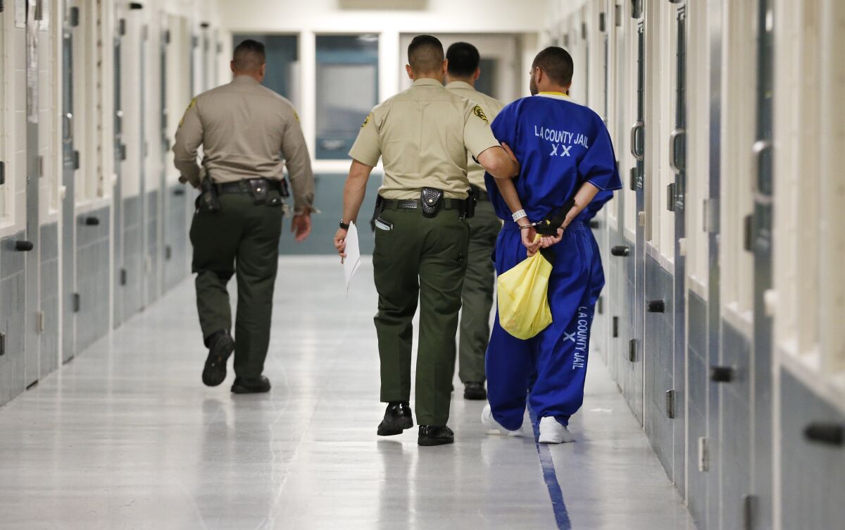 Three people in law enforcement uniforms walk with a person in an "L.A. County Jail" coverall.