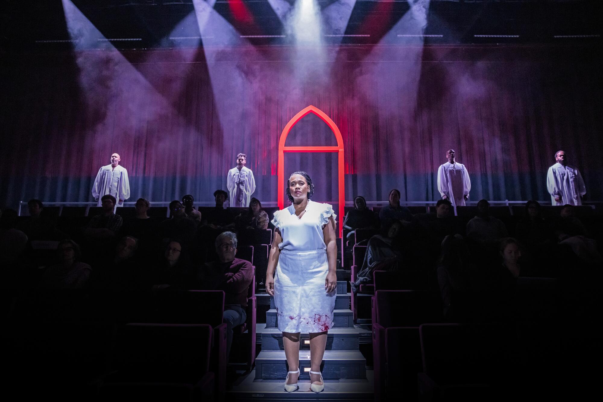 A Black woman in a blood spattered white dress stands on the steps before a stage, outlined by a red Gothic door frame