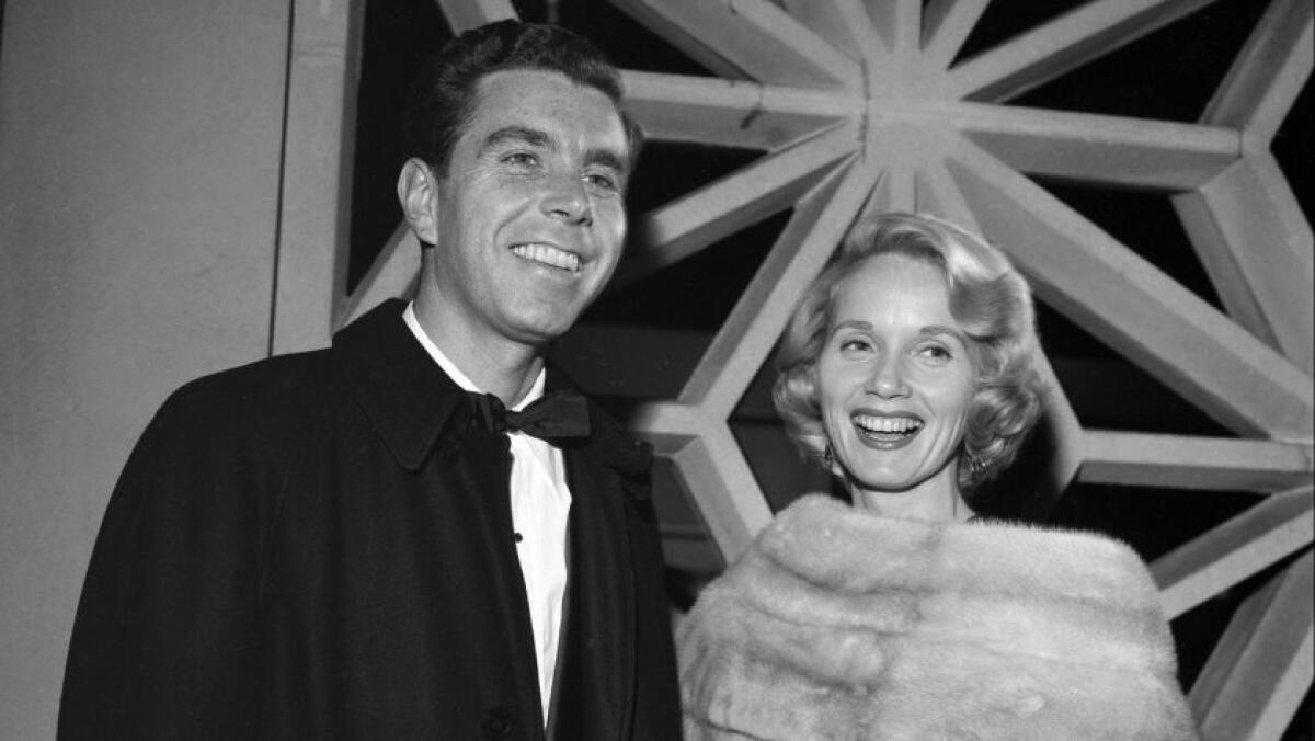 Eva Marie Saint in a fur stole and Jeffrey Hayden in a tuxedo with a bow tie stand in front of a window.