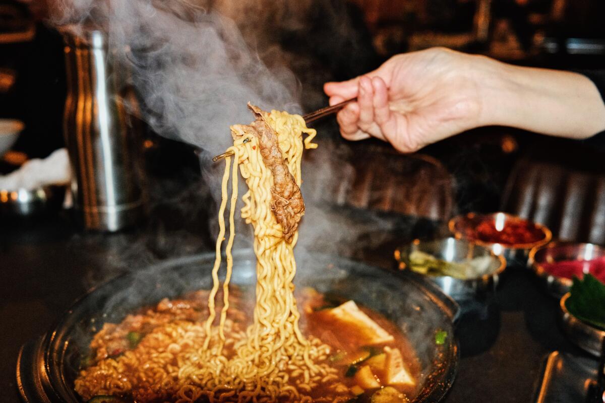 A hand with chopsticks pulling noodles and beef from a steaming bowl of stew at Origin Korean BBQ in Koreatown