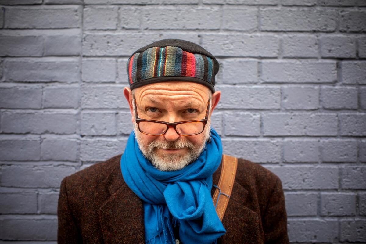 Author Francis Spufford wears a brown coat, blue scarf, glasses and colorful hat.