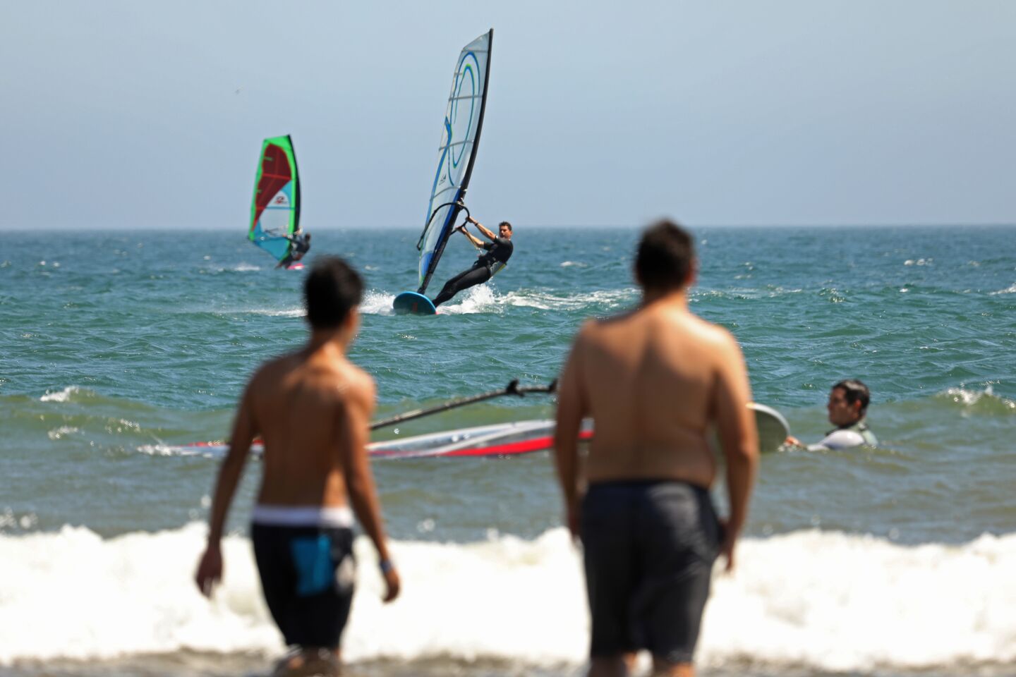 Windsurfers sail off Cabrillo Beach in San Pedro as people watch from the beach.