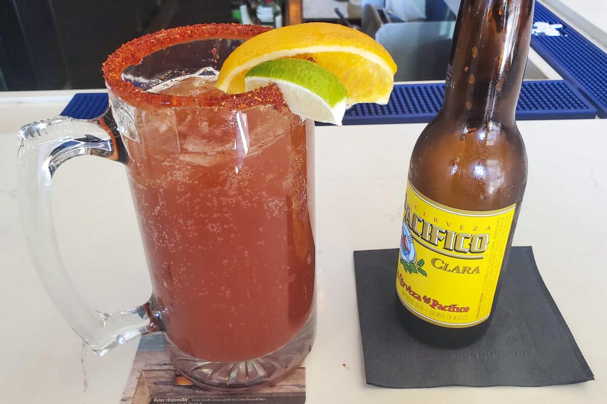 A mug full of a red michelada stands next to a bottle of Pacifico beer.