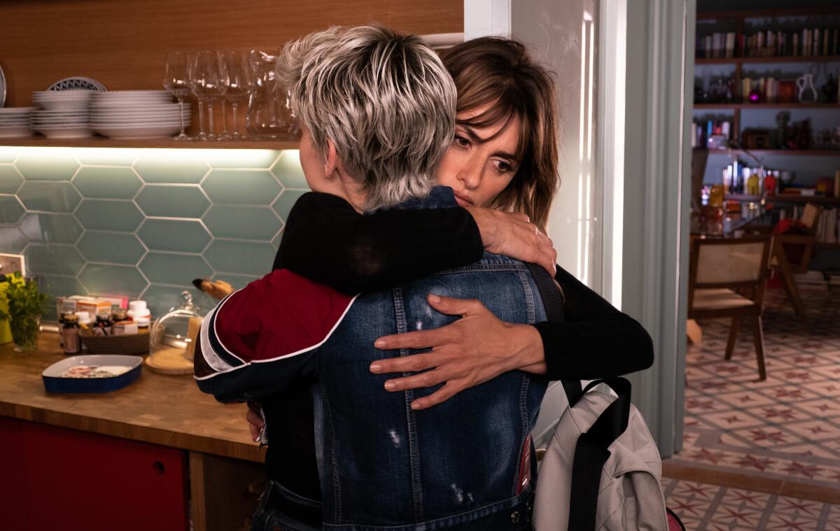 A woman with dark hair hugs a woman with short blond hair in a kitchen.