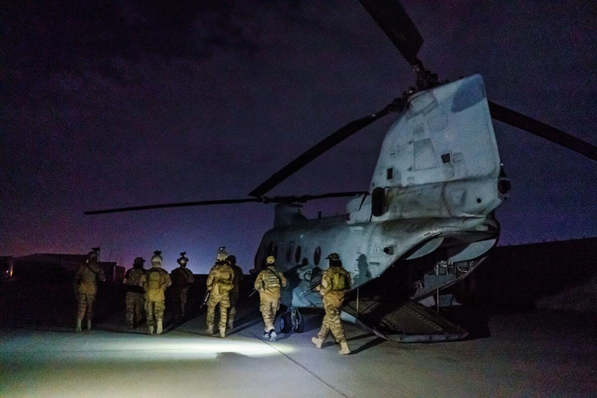 Troops in camouflage gathering by a large helicopter as night falls